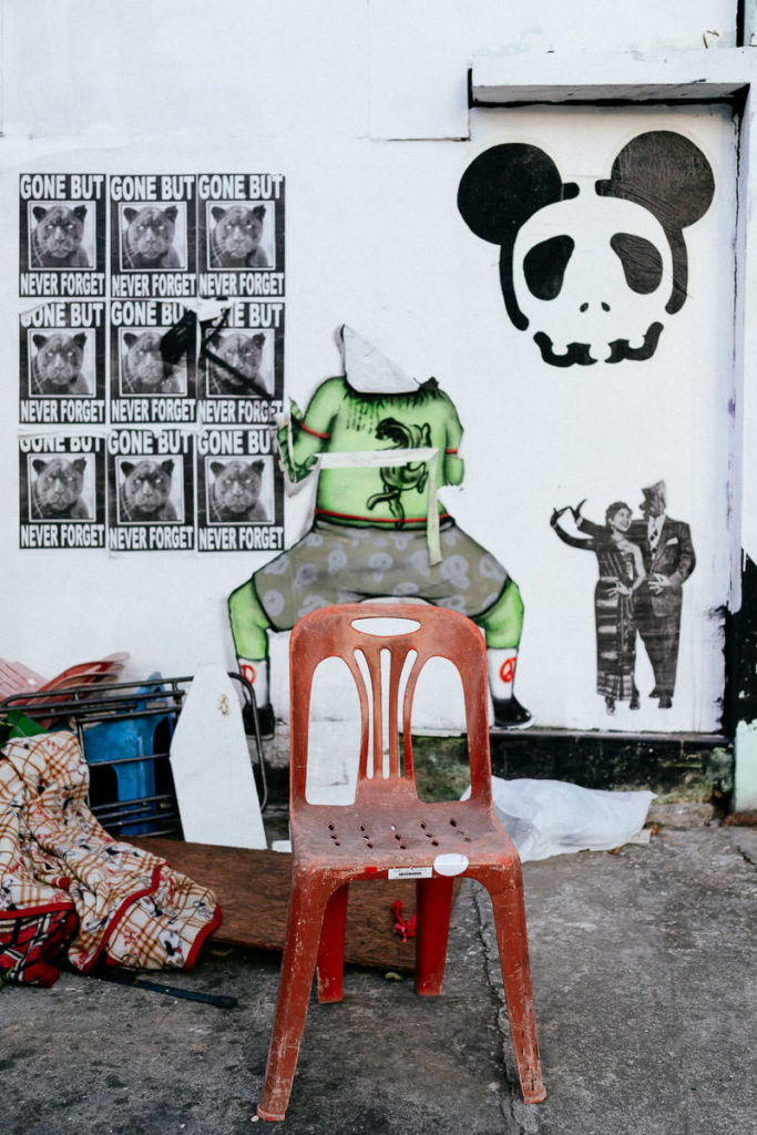 Roter Stuhl und Gerümpel vor einer Wand mit Werbung und skurrilen Graffitis